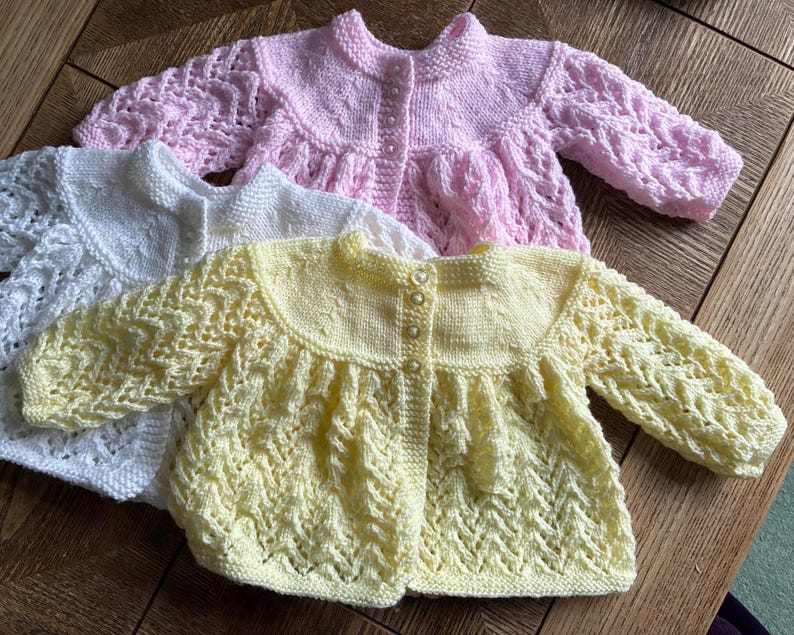 May include: Three hand-knitted baby cardigans in pastel colors: white, pink, and yellow. Each cardigan features a round collar, button closure, and intricate openwork sleeves. The garments are made of soft yarn, perfect for a baby.