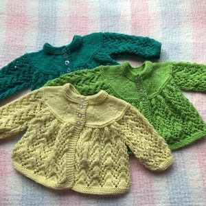 May include: Three hand-knitted baby sweaters in shades of green and yellow. The sweaters have a round collar, long sleeves, and button closures. The sweaters are made of a soft, textured yarn. The sweaters are displayed on a patterned surface.