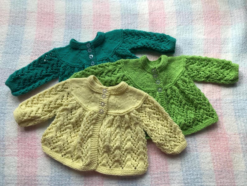 May include: Three hand-knitted baby sweaters in shades of green and yellow. The sweaters have a round collar, long sleeves, and button closures. The sweaters are made of a soft, textured yarn. The sweaters are displayed on a patterned surface.