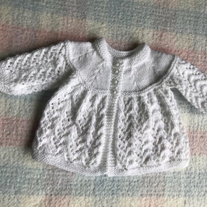 May include: A white, knitted baby cardigan with a round collar and long sleeves. The cardigan features a textured, openwork pattern on the sleeves and body, and is fastened with small, pearlescent buttons. The garment is made of a soft, white yarn.