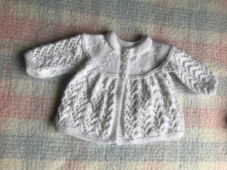 May include: A white, knitted baby cardigan with a round collar and long sleeves. The cardigan features a textured, openwork pattern on the sleeves and body, and is fastened with small, pearlescent buttons. The garment is made of a soft, white yarn.