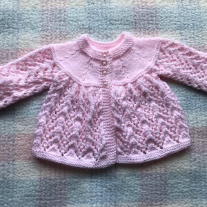 May include: A light pink, hand-knitted baby cardigan. The sweater features a round collar, long sleeves, and a button-up front. The body of the sweater has a textured, openwork design. The cardigan is displayed on a multicolored, textured surface.