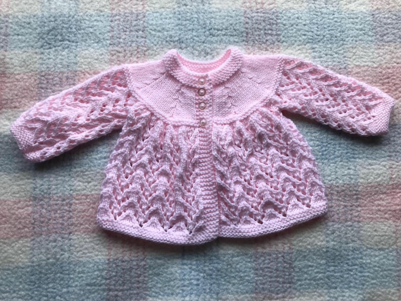 May include: A light pink, hand-knitted baby cardigan. The sweater features a round collar, long sleeves, and a button-up front. The body of the sweater has a textured, openwork design. The cardigan is displayed on a multicolored, textured surface.
