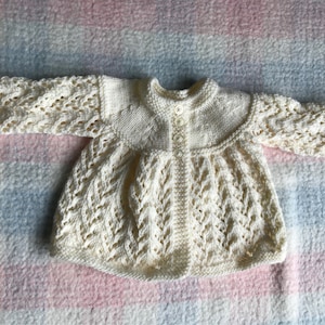 May include: Cream-colored, hand-knitted baby cardigan with a round collar and long sleeves. The cardigan features a button-up front and intricate openwork detailing on the sleeves and bodice. The garment is made of soft yarn and is designed for a baby.