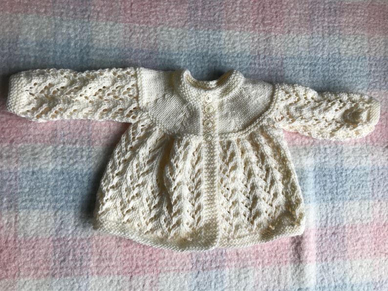 May include: Cream-colored, hand-knitted baby cardigan with a round collar and long sleeves. The cardigan features a button-up front and intricate openwork detailing on the sleeves and bodice. The garment is made of soft yarn and is designed for a baby.