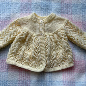May include: A cream-colored, hand-knitted baby cardigan with a round collar and button closure. The sleeves and body feature an openwork design. The garment is displayed on a pink and blue plaid blanket.
