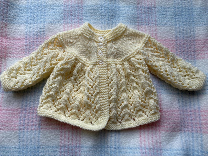 May include: A cream-colored, hand-knitted baby cardigan with a round collar and button closure. The sleeves and body feature an openwork design. The garment is displayed on a pink and blue plaid blanket.