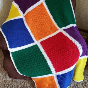 May include: A colorful crochet blanket with a geometric pattern of squares in purple, yellow, orange, blue, green, and red. The squares are outlined in white. The blanket is draped over a brown chair.