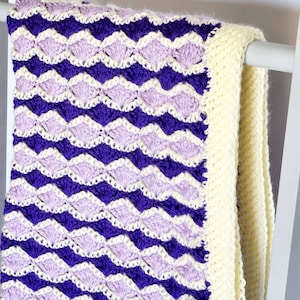 May include: A crocheted baby blanket with a purple and lavender chevron pattern and a white border.