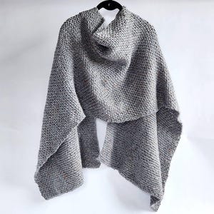 Puede incluir: Un chal gris tejido a mano con una apariencia texturizada, colgado de una percha negra. El chal está hecho de un hilo suave y moteado, con un diseño de cuello alto. Un accesorio acogedor para el clima fresco.