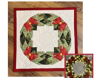 May include: A quilted Christmas wreath wall hanging with a red border and a green and red wreath design. The wreath is made of fabric and has a white background.