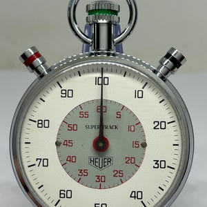 Op de afbeelding: Een vintage Heuer Super Track stopwatch met een witte wijzerplaat, zwarte cijfers en rode accenten. De stopwatch heeft een chromen behuizing en een centrale wijzerplaat met het Heuer-logo. Het horloge is gemarkeerd met "Made in Switzerland".