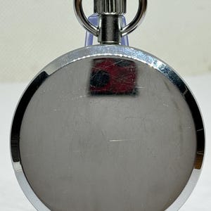 May include: A silver-colored pocket watch with a circular face and a metal loop at the top. The watch has a shiny, reflective surface with a textured winding stem. The watch is displayed on a clear plastic stand.