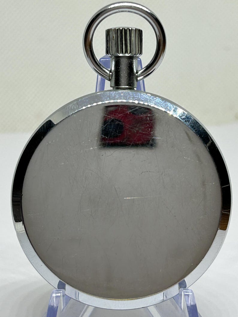 May include: A silver-colored pocket watch with a circular face and a metal loop at the top. The watch has a shiny, reflective surface with a textured winding stem. The watch is displayed on a clear plastic stand.