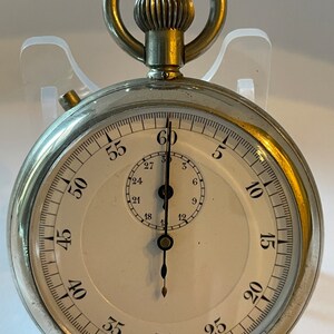 Puede incluir: Un reloj de bolsillo plateado con una esfera blanca y marcas negras. El reloj tiene una aguja grande para los segundos y una aguja más pequeña para los minutos. La esfera del reloj tiene números del 5 al 60 alrededor del borde. El reloj tiene una cadena plateada unida a la parte superior.