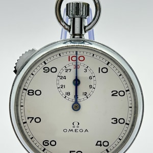 May include: A silver-toned Omega stopwatch with a white dial, featuring black numerals from 10 to 100, a smaller sub-dial with numerals 3 to 27, a blue central hand, a top winding crown with a circular ring, and the text OMEGA and SWISS MADE.