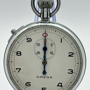 Puede incluir: Un cronómetro Omega de tono plateado con esfera blanca, números arábigos negros del uno al nueve y una subesfera central. El reloj presenta manecillas azules, un marcador circular rojo en la parte superior, una corona de cuerda y el texto OMEGA y SWISS MADE.