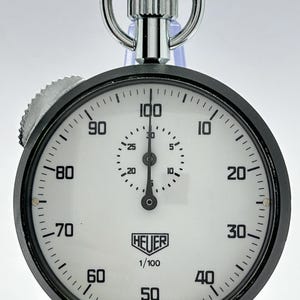 May include: A vintage Heuer stopwatch with a black circular case and a white dial. The face features black numerals from 10 to 100, a central sub-dial, and the text Heuer 1/100 Made in Switzerland. It includes a silver crown and a top ring.