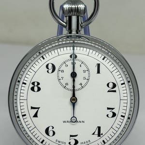 May include: A silver-toned pocket watch with a white face, featuring black Arabic numerals and a smaller subdial. The watch has a ring at the top and the words "WARMANN" and "SWISS MADE" are visible.