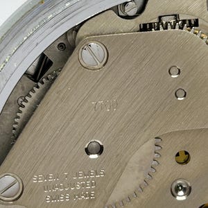 May include: Close-up of a watch movement with visible gears and components. The metal plate is marked with "7700" and "SEVEN 7 JEWELS UNADJUSTED SWISS MADE". The watch mechanism is silver-toned and detailed.