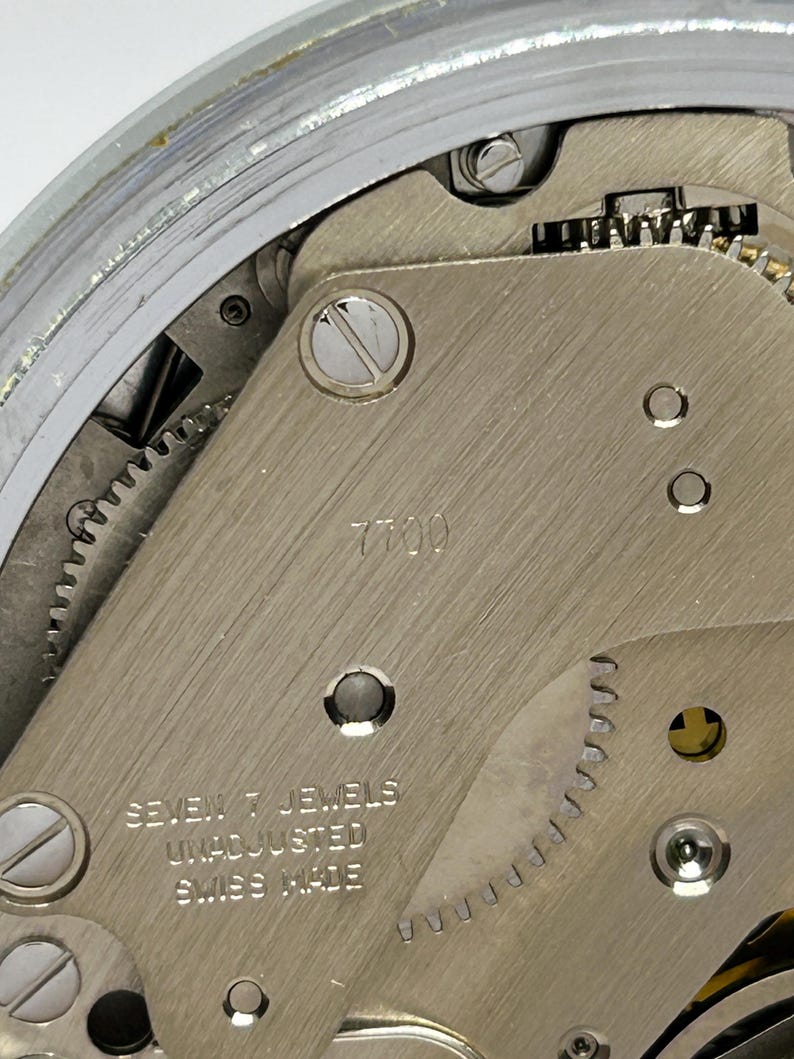 May include: Close-up of a watch movement with visible gears and components. The metal plate is marked with "7700" and "SEVEN 7 JEWELS UNADJUSTED SWISS MADE". The watch mechanism is silver-toned and detailed.