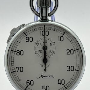 Op de afbeelding: Een zilverkleurige analoge stopwatch met een witte wijzerplaat, zwarte cijfers van 10 tot 100 en een kleinere sub-wijzerplaat van 5 tot 30. De wijzerplaat bevat de tekst Minerva en Patent. Inclusief een opwindkroon met een ronde ring.