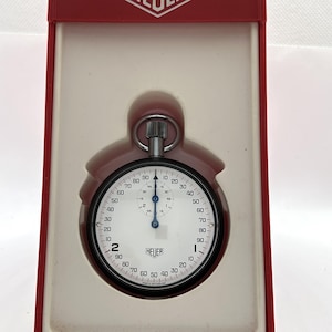 Op de afbeelding: Een Heuer stopwatch met een witte wijzerplaat, zwarte cijfers en blauwe wijzers, geplaatst in een rood en crèmekleurige kunststof displaydoos. De stopwatch heeft een opwindkroon aan de bovenkant en het Heuer-logo op de wijzerplaat en de deksel.