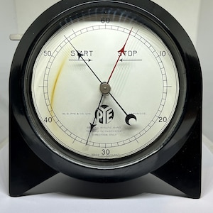 Peut inclure: Un chronomètre noir vintage avec un cadran blanc et des aiguilles noires. Le cadran circulaire affiche des graduations de 0 à 60. Les mots "START" et "STOP" sont visibles. Le texte "W.G. PYE & CO. LTD" et "MADE IN ENGLAND" sont également présents.