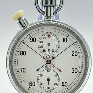 May include: A silver-toned Lemania Nero stopwatch with a white dial, featuring black numerals from 10 to 90, a red hand, a blue hand, and two smaller sub-dials. The top includes a winding crown and a white pusher button, with Swiss Made text.