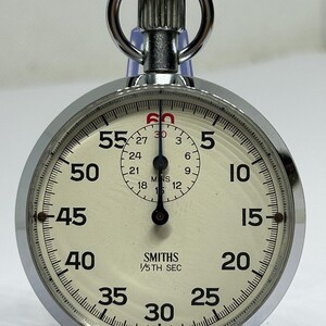May include: A vintage Smiths stopwatch with a silver-colored metal casing and a cream-colored face. The watch features black numerals and markings, with a smaller dial in the center. The text "SMITHS 1/5TH SEC" is printed on the face.
