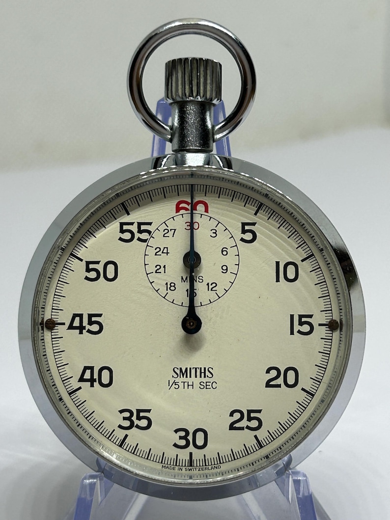 May include: A vintage Smiths stopwatch with a silver-colored metal casing and a cream-colored face. The watch features black numerals and markings, with a smaller dial in the center. The text "SMITHS 1/5TH SEC" is printed on the face.