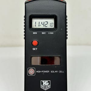 May include: Black TAG Heuer Microsplit 200 SOLAR stopwatch with a digital display showing 11:42:10. It has red buttons for SPL-RESET and START/STOP, and a solar cell. The stopwatch is designed for timing events.