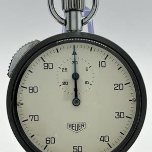 Może przedstawiać: Zabytkowy stoper Heuer z okrągłą czarną metalową kopertą i kremową tarczą. Tarcza posiada czarne cyfry od 10 do 90, centralną niebieską wskazówkę, małą tarczę pomocniczą oraz napis Heuer i Made in Switzerland.