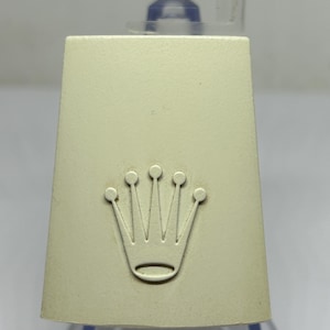 Può includere: Scatola rettangolare color crema con il logo della corona Rolex in rilievo sul davanti. La corona è un emblema stilizzato con cinque punte e una base curva. La scatola ha una forma leggermente trapezoidale.