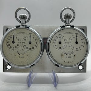 May include: Two silver-toned Junghans Process Time Meter stopwatches mounted on a rectangular metal plate. Each watch features a cream-coloured face with two main dials for minutes and seconds, a smaller 1/10 second sub-dial, and a top winding crown.