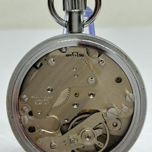 May include: A silver-toned pocket watch with a visible mechanical movement. The watch features a circular face with intricate gears, screws, and a balance wheel. The watch has a winding stem and a loop for a chain. The text "SEVEN 7 JEWELS UNADJUSTED SWISS MADE" is visible.