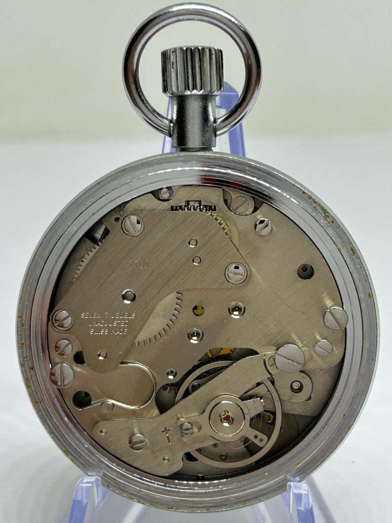 May include: A silver-toned pocket watch with a visible mechanical movement. The watch features a circular face with intricate gears, screws, and a balance wheel. The watch has a winding stem and a loop for a chain. The text "SEVEN 7 JEWELS UNADJUSTED SWISS MADE" is visible.