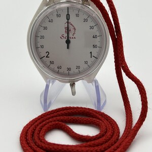 May include: A silver-toned analog stopwatch with a white face, black numerals, and a single black hand. The dial features the text "Sirius" and a small graphic of a person in a headdress. A thick red braided cord is attached to the top.
