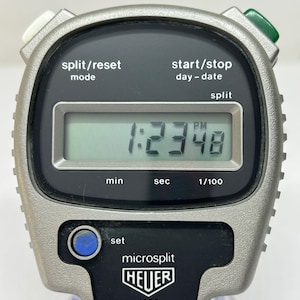 May include: A silver Heuer microsplit stopwatch displaying 1:23:48 PM. The stopwatch has black buttons labeled "split/reset" and "start/stop". The brand name "HEUER" is visible, along with the model "1000".