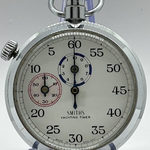 Peut inclure: Un chronomètre de yachting Smiths argenté avec un cadran blanc, des chiffres noirs et une couronne de remontage sur le dessus. Le cadran présente un sous-cadran bleu pour les minutes et un sous-cadran à bordure rouge pour les secondes.