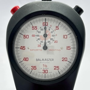 May include: A black Balmaster Antichoc stopwatch with a white face, red numerals, and a green-accented crown. The dial features markings for Sports and Yachting, 4 rubis, and 1/5 second increments. It includes a red button and a black button.