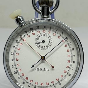May include: A vintage Lemania stopwatch with a white face, black and red hands, and a chrome casing. The watch has a small seconds dial and a crown. The face displays numbers from 1 to 60, with the text "SWISS MADE".