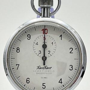 May include: A silver-tone Hanhart stopwatch with a white dial, black numerals from 1 to 10, and a smaller sub-dial. The face features the text Hanhart, LEVER 7 JEWELS, SHOCK PROOF, 1/10 SEC, and MADE IN GERMANY. It has a circular metal casing.