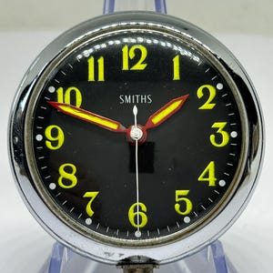 Może przedstawiać: Zegar Smiths w stylu vintage z czarną tarczą, żółtymi cyframi i żółto-czerwonymi wskazówkami. Zegar ma chromowaną ramkę i trzpień do nakręcania. Zegar jest wystawiony na przezroczystym plastikowym stojaku.