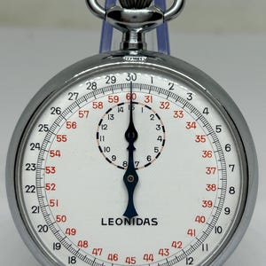 May include: A classic stopwatch with a white dial and black numerals. The stopwatch features a silver metal casing and a crown. The brand name "LEONIDAS" is printed on the face. The stopwatch is displayed on a clear stand.