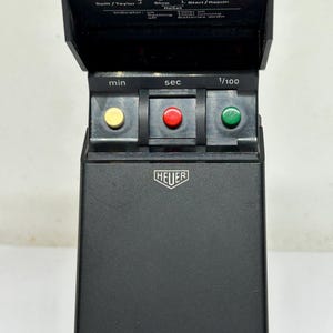 May include: A black Heuer stopwatch with a flip-up cover. The stopwatch has three buttons: yellow, red, and green. The face displays "min", "sec", and "1/100". The Heuer logo is visible on the front.