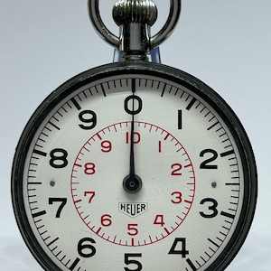 Può includere: Un cronometro Heuer vintage con quadrante bianco, numeri neri e indicatori interni rossi. Il cronometro ha una cassa in metallo, una corona di carica e un anello per appenderlo. Il testo "HEUER" è visibile. È contrassegnato "MADE IN SWITZERLAND".
