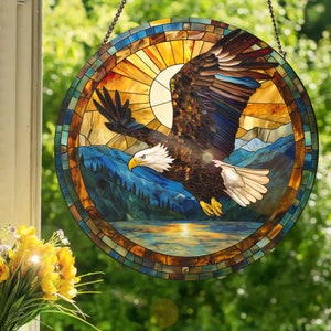 Könnte beinhalten: Ein Buntglasfenster, das einen Weißkopfseeadler zeigt, der über einem Gebirge mit einem See im Vordergrund fliegt. Der Adler ist braun und weiß mit einem gelben Schnabel. Die Berge sind blau und grün. Der See ist blau mit einem Sonnenuntergang im Hintergrund.