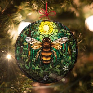 May include: Round Christmas ornament featuring a stained-glass design of a bee in a forest setting. The bee has brown and yellow stripes, with white and yellow wings. The background includes green foliage, trees, and a yellow sun. A red ribbon is attached for hanging.