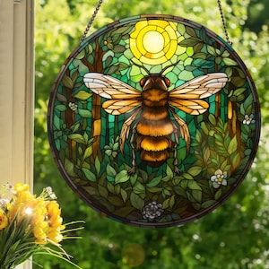 May include: Round stained glass window art featuring a detailed bee with outstretched wings, set against a forest scene with a yellow sun. The artwork uses shades of green, brown, and yellow, and is suspended by a chain.